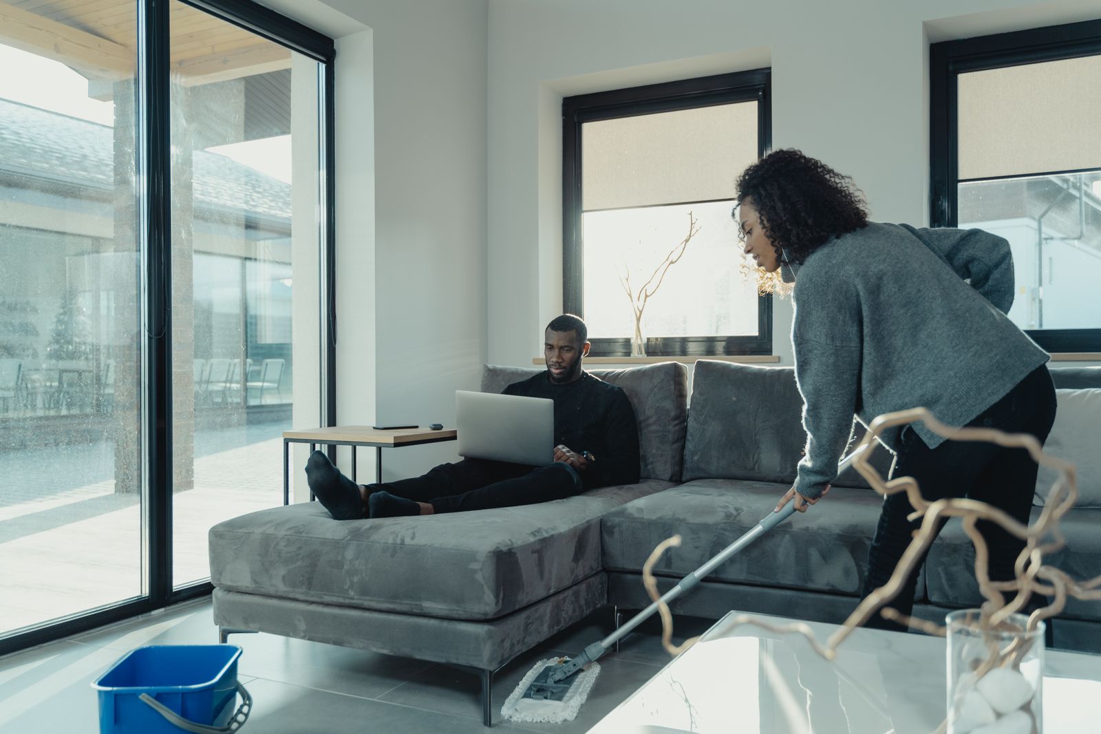 Eine Frau wischt den Boden im Wohnzimmer, während ein Mann auf dem Sofa sitzt